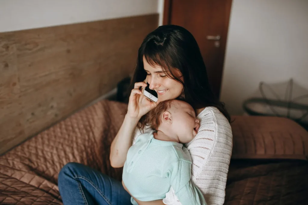 Smiling mom on a phone call holding her sleeping baby on a bed