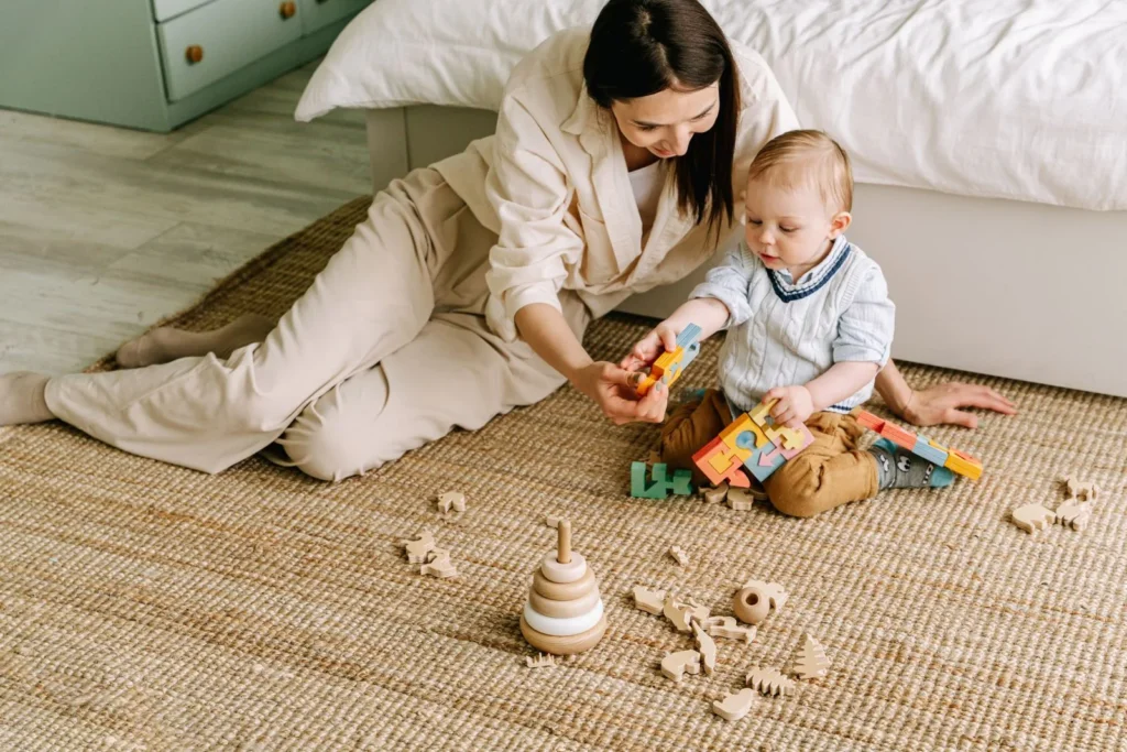 Caregiver on a woven rug playing with a baby and wooden stacking and block toys