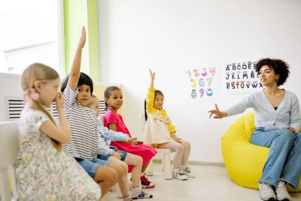 Five pre-K-age children in colorful clothes raising hands to a teacher seated on a yellow bean bag, alphabet posters on the wall