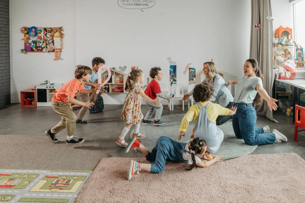 Two caregivers on the floor with a group of preschoolers playing and running in a bright childcare room with wall art
