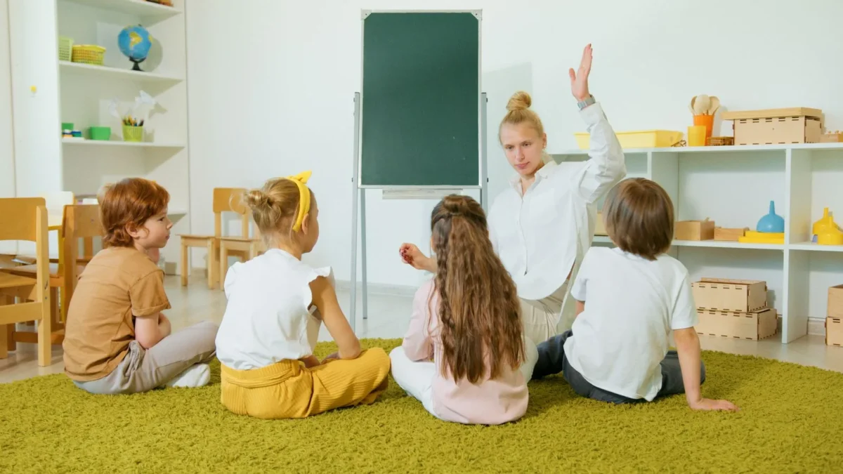 Teacher with one hand raised leading four seated children on a green carpet in a bright classroom with shelves and a green board