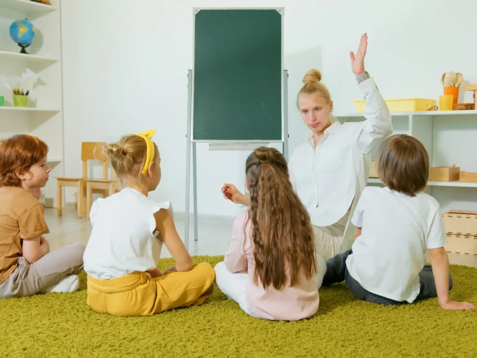 Teacher with one hand raised leading four seated children on a green carpet in a bright classroom with shelves and a green board